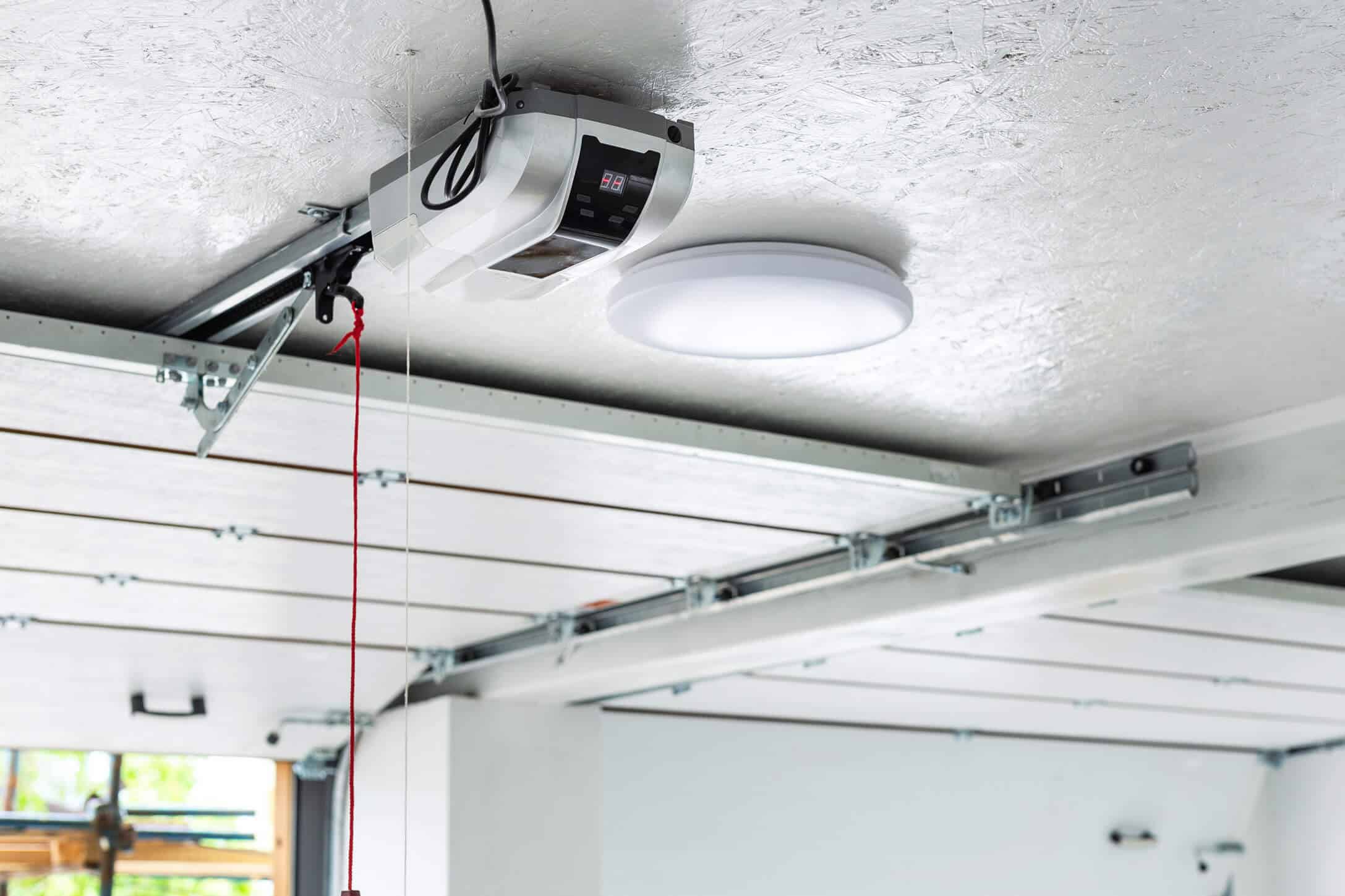 A close-up view of a garage ceiling features a mounted garage door opener—essential for those who want to Know About Garage Door Openers—with a power cord, red pull handle, round ceiling light, and visible metal tracks.