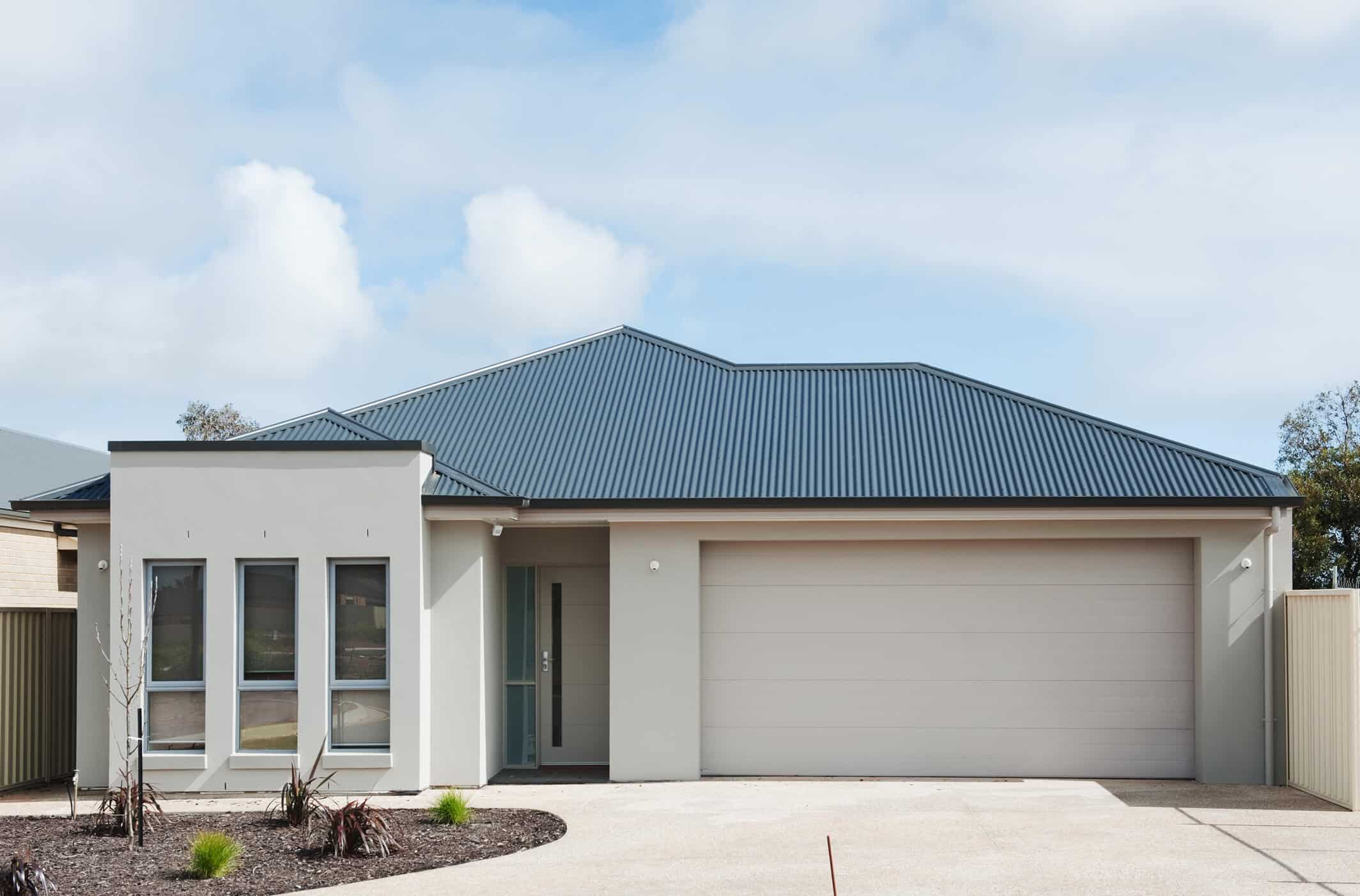 Modern single-story house with a gray metal roof, light-colored exterior walls, three vertical windows on the left, a glass front door in the center, and sleek contemporary garage doors on the right. Simple landscaping in front.