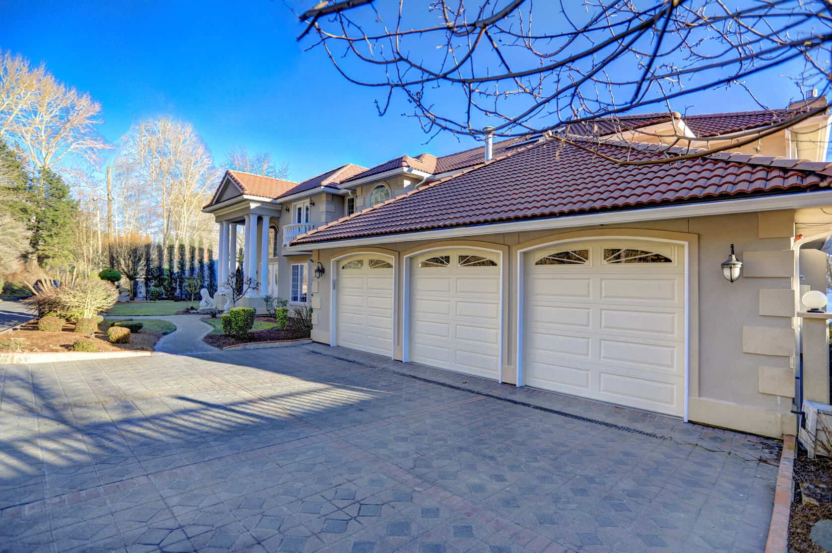 Large, elegant house with a tile roof and three-car garage—perfect if you’ve ever thought, “Not enough space in the garage?” Manicured landscaping frames the entrance, with a stone driveway and clear blue sky overhead.