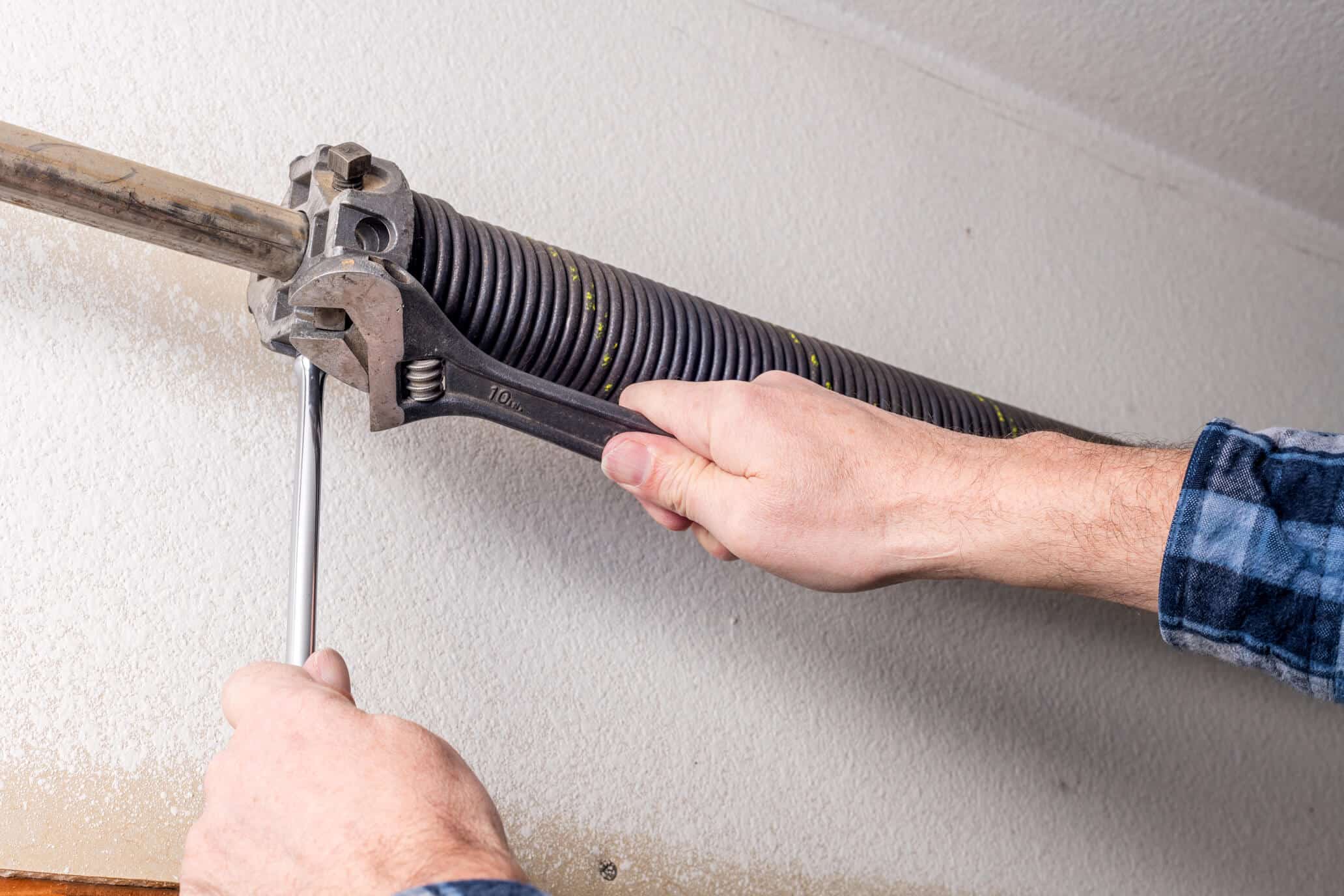 A person uses a wrench and a metal rod to adjust or repair a black torsion spring on a garage door mechanism mounted on a wall, raising the question: Should I repair my garage door myself?.