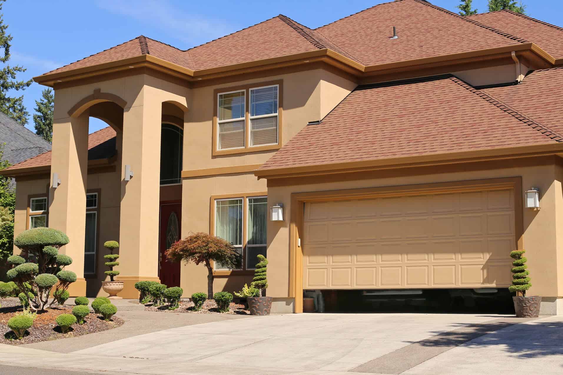 Two-story tan stucco house with a brown shingle roof, double garage—ideal for those asking, "Which Garage Door Is Best for Me?" Large windows and front yard landscaping with trimmed bushes and a small tree complete the look on this bright sunny day.