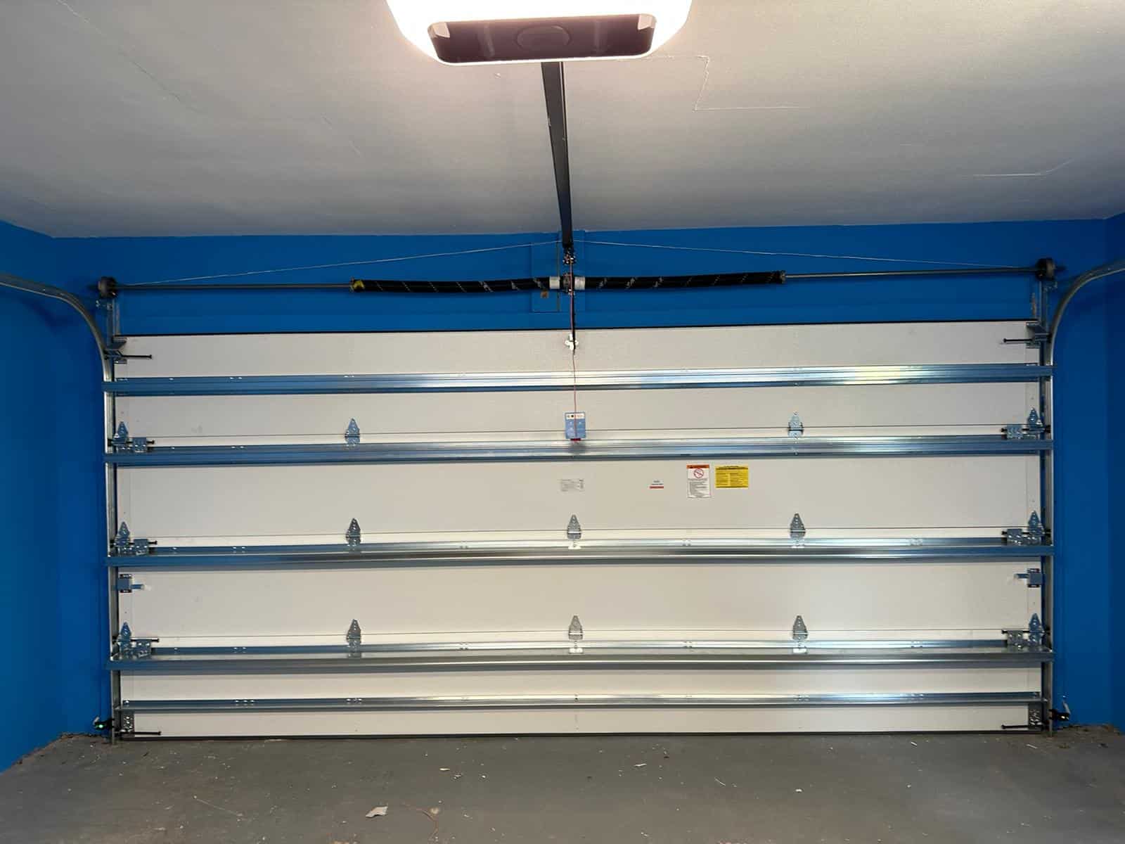 A closed white garage door with horizontal metal reinforcement bars, inside a garage with bright blue walls and a gray floor. Overhead garage storage is installed above the door, maximizing storage space without crowding the area.