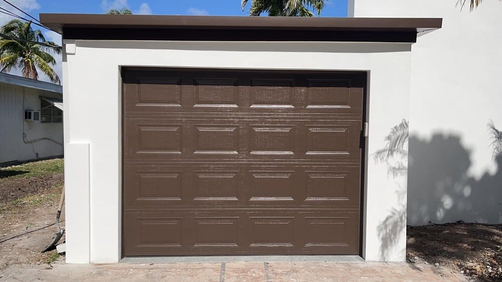A small, single-car garage with a dark brown, paneled garage door and white exterior walls—a perfect spot for a DIY makeover—sits next to a house surrounded by palm trees and blue sky in the background.