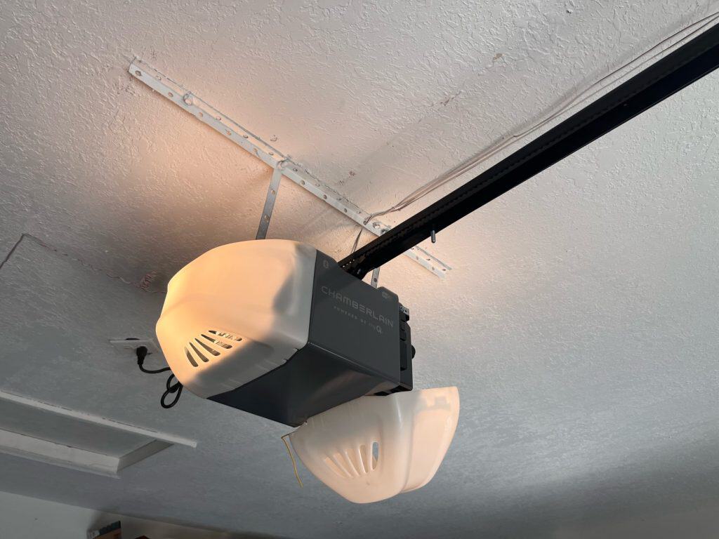 Ceiling-mounted garage door opener with two white plastic light covers, one hanging loose. This device, a common example among types of garage door openers, is attached to a black rail beneath a textured white ceiling.