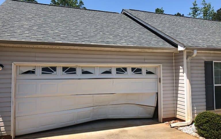 A white garage door with rectangular windows is bent and dented along the bottom and middle panels, unlike sturdy hurricane rated doors that protect your home from severe weather. The damage creates a warped appearance on a light-colored house.