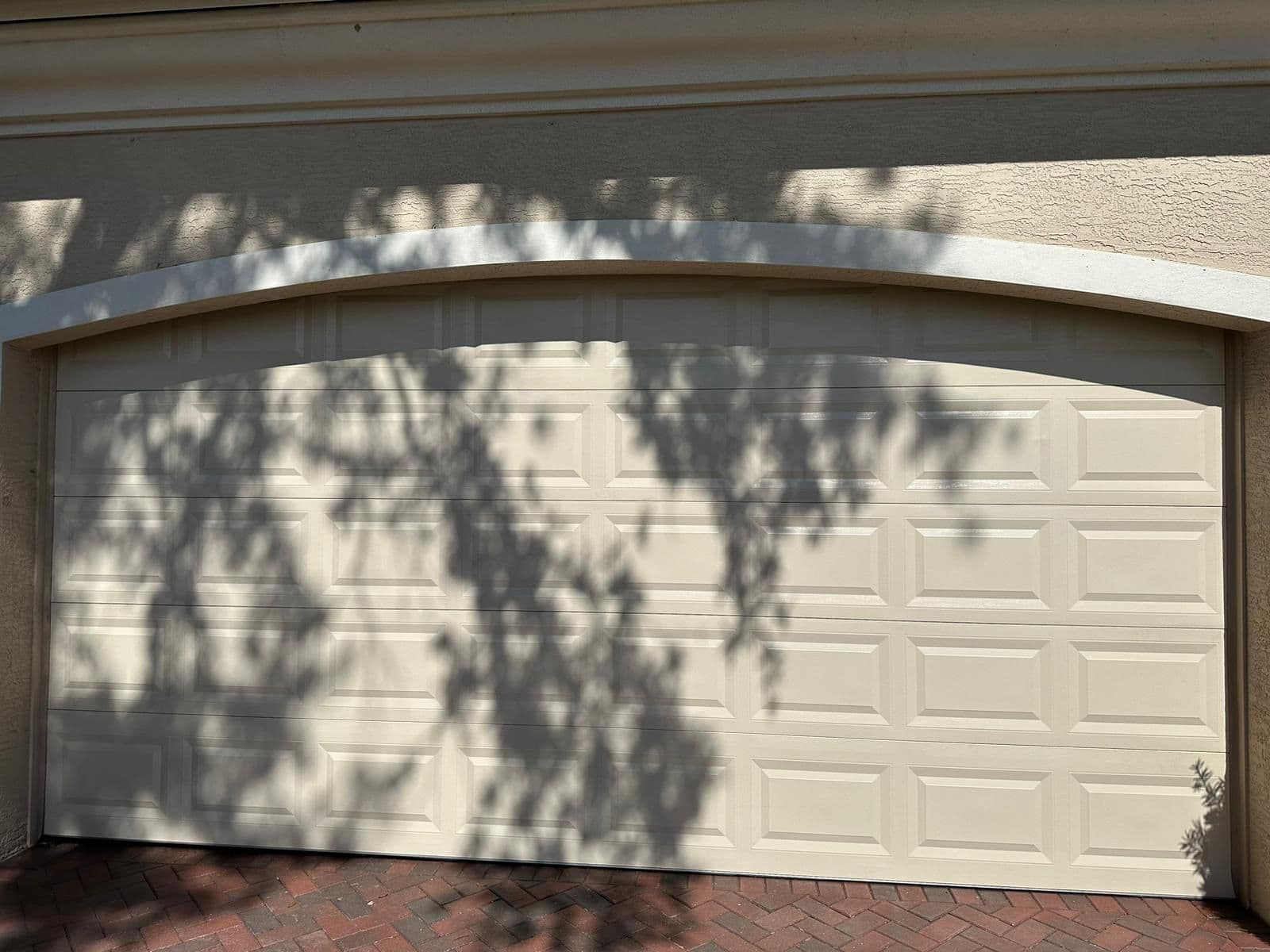 A closed beige garage door with a rectangular panel design, partially shaded by tree branches casting distinct shadows, offers added coverage for your valuables above a red brick driveway.