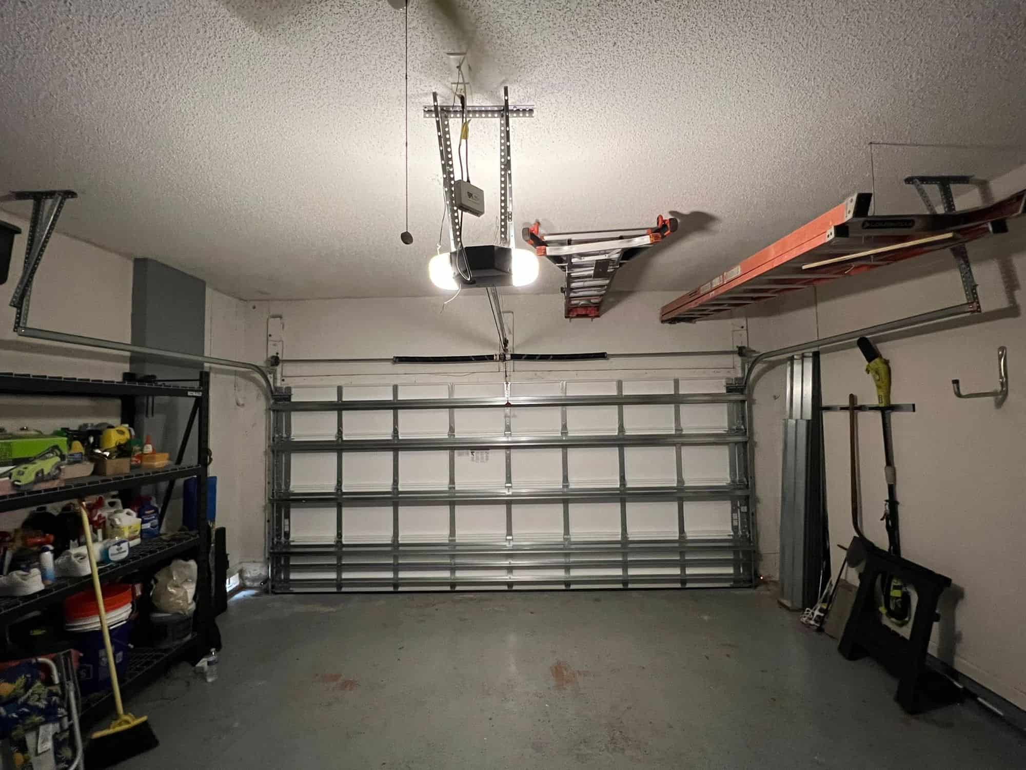 A clean, empty garage with a closed metal garage door highlights the benefits of professional garage door service, featuring ceiling lights, shelves on the left, ladders overhead, and tools neatly stored against the right wall.