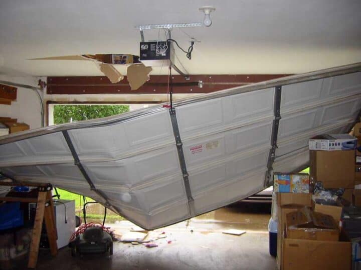 A garage door is partially detached and hangs at an angle from the ceiling, with visible damage and insulation above—a clear reminder of the importance of home safety during hurricane season. Boxes and tools are scattered around the cluttered garage.