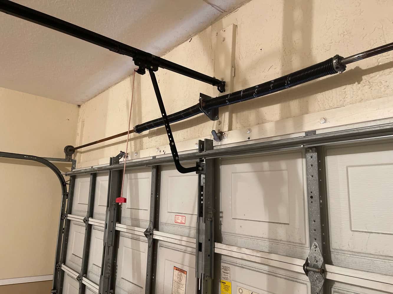 Garage door interior showing metal tracks, a torsion spring, and support brackets above the door. A red cord hangs for manual operation. Proper garage spring repair is crucial due to garage spring risks. The wall and ceiling are beige.