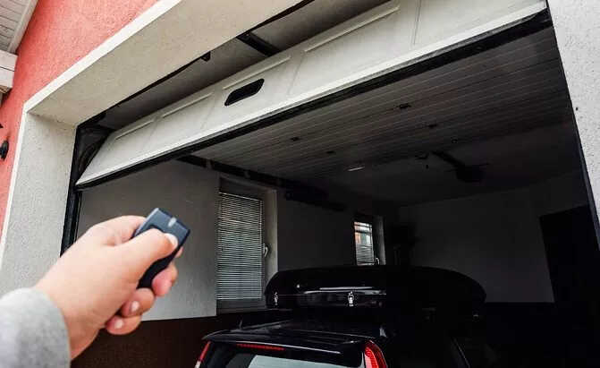 A person holds a remote control to open a partially raised garage door, revealing a black car parked inside a garage attached to a house with a red exterior wall—a common scene for users of Garage Door Openers in South Florida.