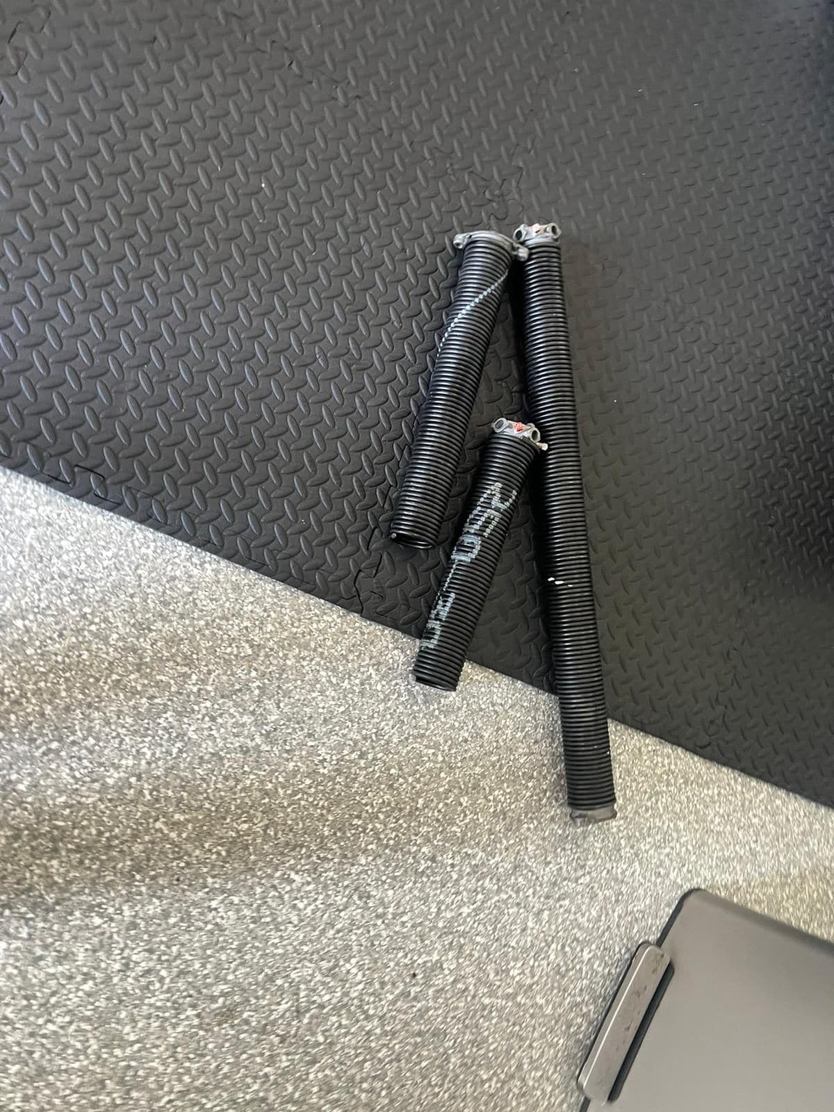 Three black metal garage door torsion springs with colored ends lie on a floor that transitions from gray speckled carpet to black rubber matting, illustrating part of the garage door repair process. A portion of a gray object appears in the bottom right corner.