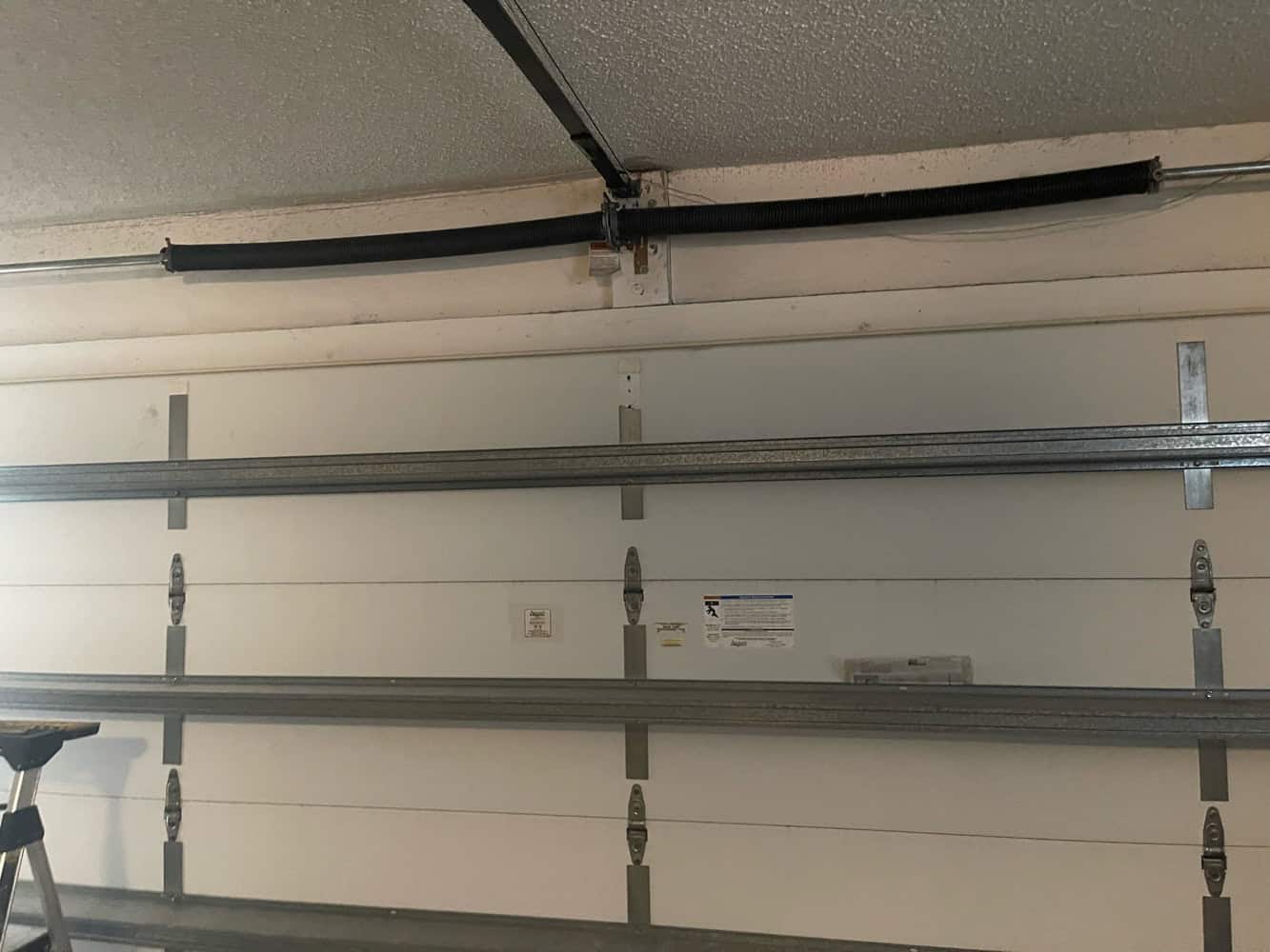 A white garage door with metal horizontal supports and a clearly broken torsion spring above it. The black spring is split in the middle, making you wonder: Who Do I Call For Garage Door Spring Repair? The garage door remains closed.