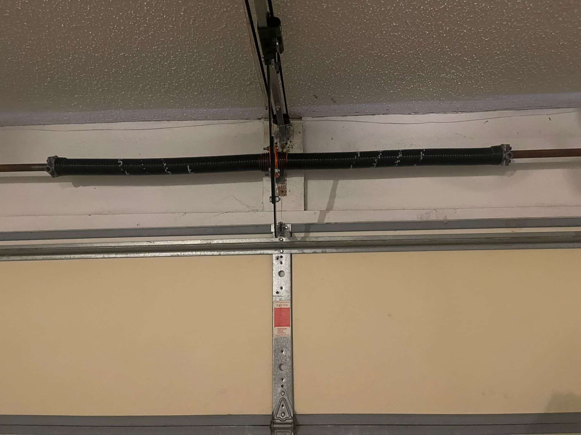 A close-up view of a garage door torsion spring mounted above a closed garage door, attached to a metal bar and bracket on the ceiling. The intact spring ensures smooth functionality and security for your garage.