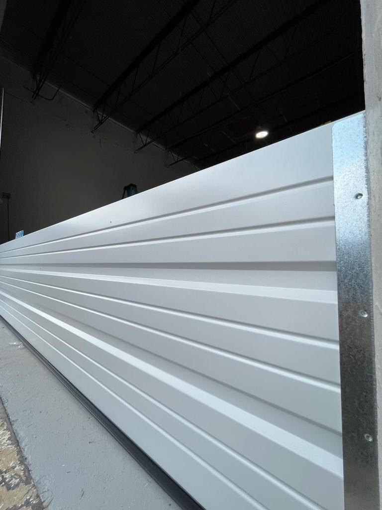 A close-up view of a large, white, horizontally-ridged metal door with garage door panels, partially open inside a spacious industrial or warehouse setting with a high ceiling and exposed beams, blending functionality with aesthetic appeal.