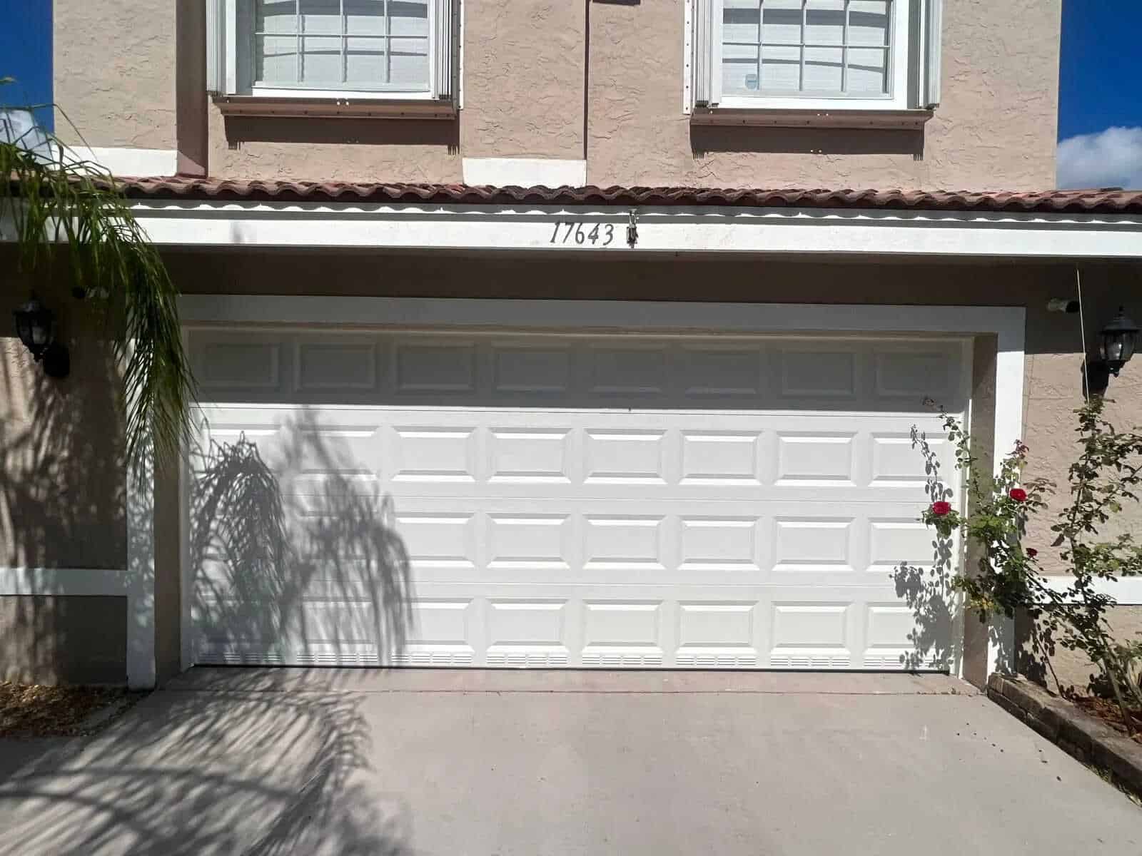 A closed, painted white garage door is attached to a beige two-story home exterior with tile roofing. Shadows from nearby plants fall on the driveway, and a few rose bushes grow beside the garage. The house number 17643 is visible above the door.