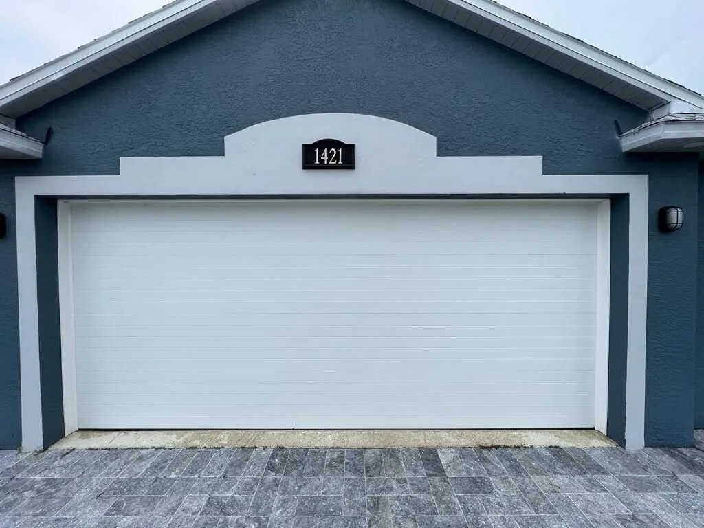 A closed white magnetic garage door on a blue-gray house, with a plaque above displaying the number 1421. The driveway is paved with gray tiles.