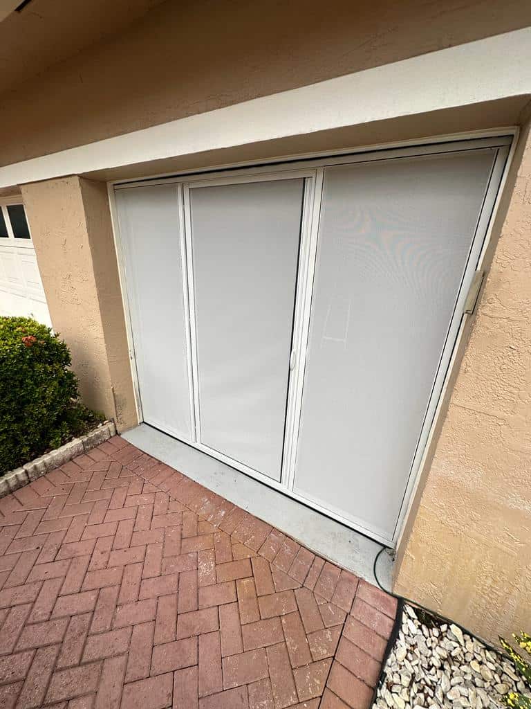 Sliding glass door covered with white opaque panels, set in a beige exterior wall next to greenery and a red brick walkway. White rocks border the walkway's corner, complementing nearby Garage Door Screens for added privacy and style.