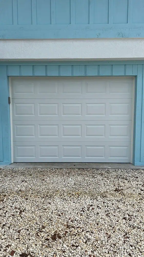 A closed white garage door is framed by light blue trim in Key Largo, FL. The ground in front is covered with small white stones. The building’s exterior is also painted light blue with a white section above the door.