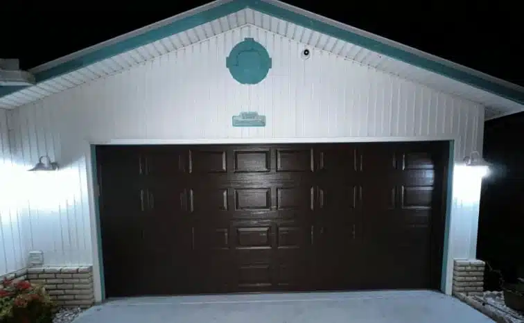 A closed, dark brown garage door on a white Fort Myers house with brick accents, lit by two bright wall-mounted lights on either side at night.
