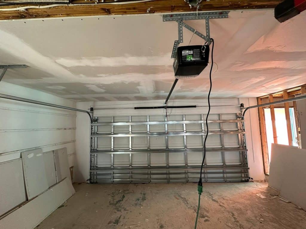 A partially finished garage with an overhead garage door, exposed ceiling beams, a garage door opener, and construction materials and panels leaning against the wall. Light enters through an unfinished window.