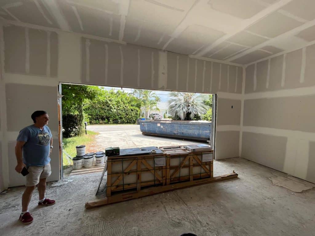 A man in a blue shirt and red shoes stands inside a partially finished Fort Lauderdale garage with unpainted drywall. The open door reveals a driveway, trees, and a large blue dumpster outside, with construction materials stacked inside.