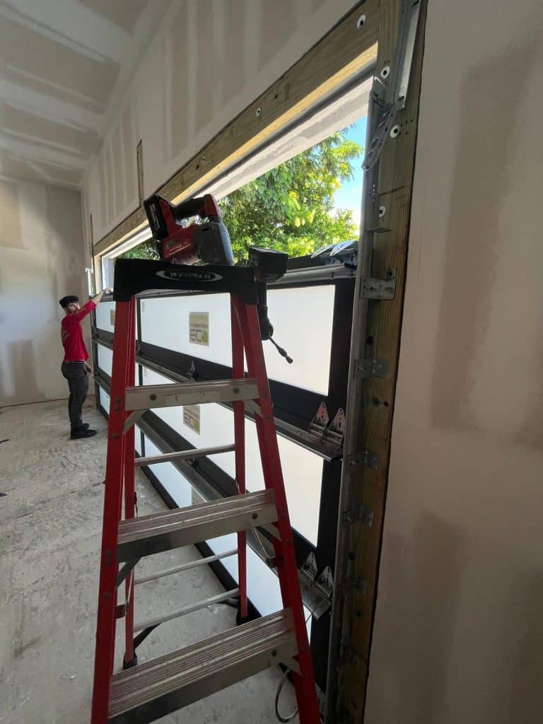 A red ladder stands in front of a partially installed garage door in a room under construction in Fort Lauderdale, FL. A person in a red shirt works near the open section, with green trees visible outside through the opening.