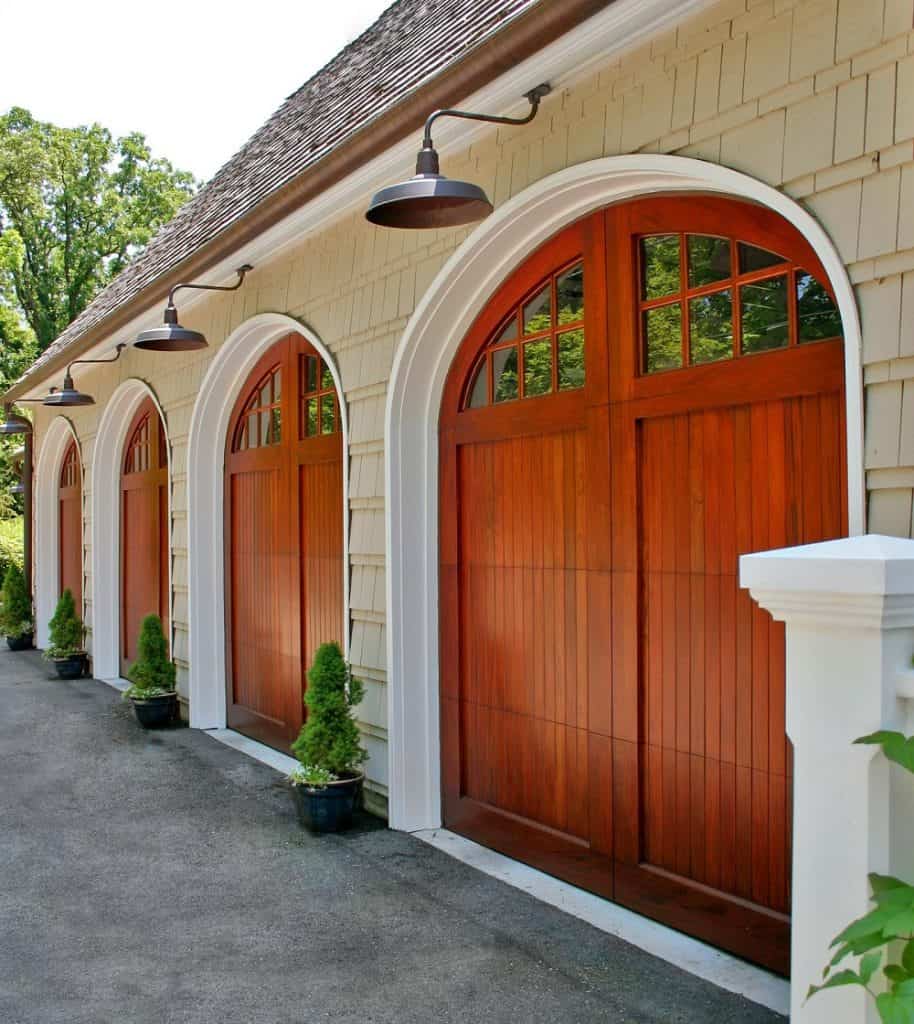 Three arched Cambek garage doors with upper windows, framed in white trim, are set in a light-colored exterior wall. Small potted shrubs line the driveway, and black gooseneck lights are mounted above each door.