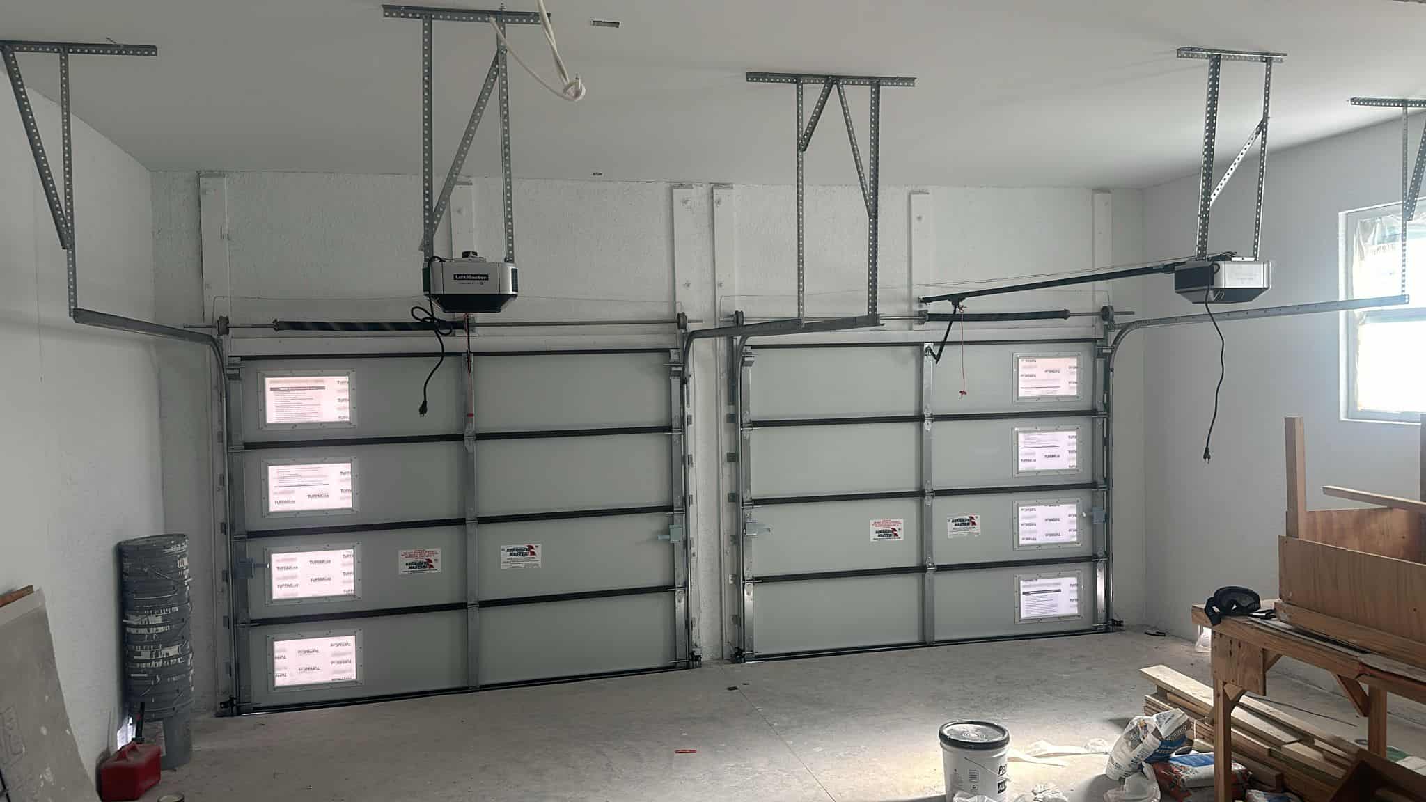 A garage interior in Naples, Florida 34102 features two closed, light gray garage doors with ceiling-mounted automatic openers. Construction materials and tools are scattered on the unfinished concrete floor as sunlight enters through small windows.
