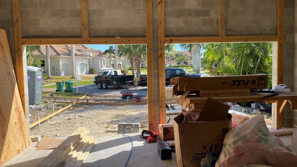 View from inside a house under construction in Naples, Florida 34102, looking out through a large window opening. Outside, see parked trucks, a portable toilet, palm trees, and houses in this suburban neighborhood.