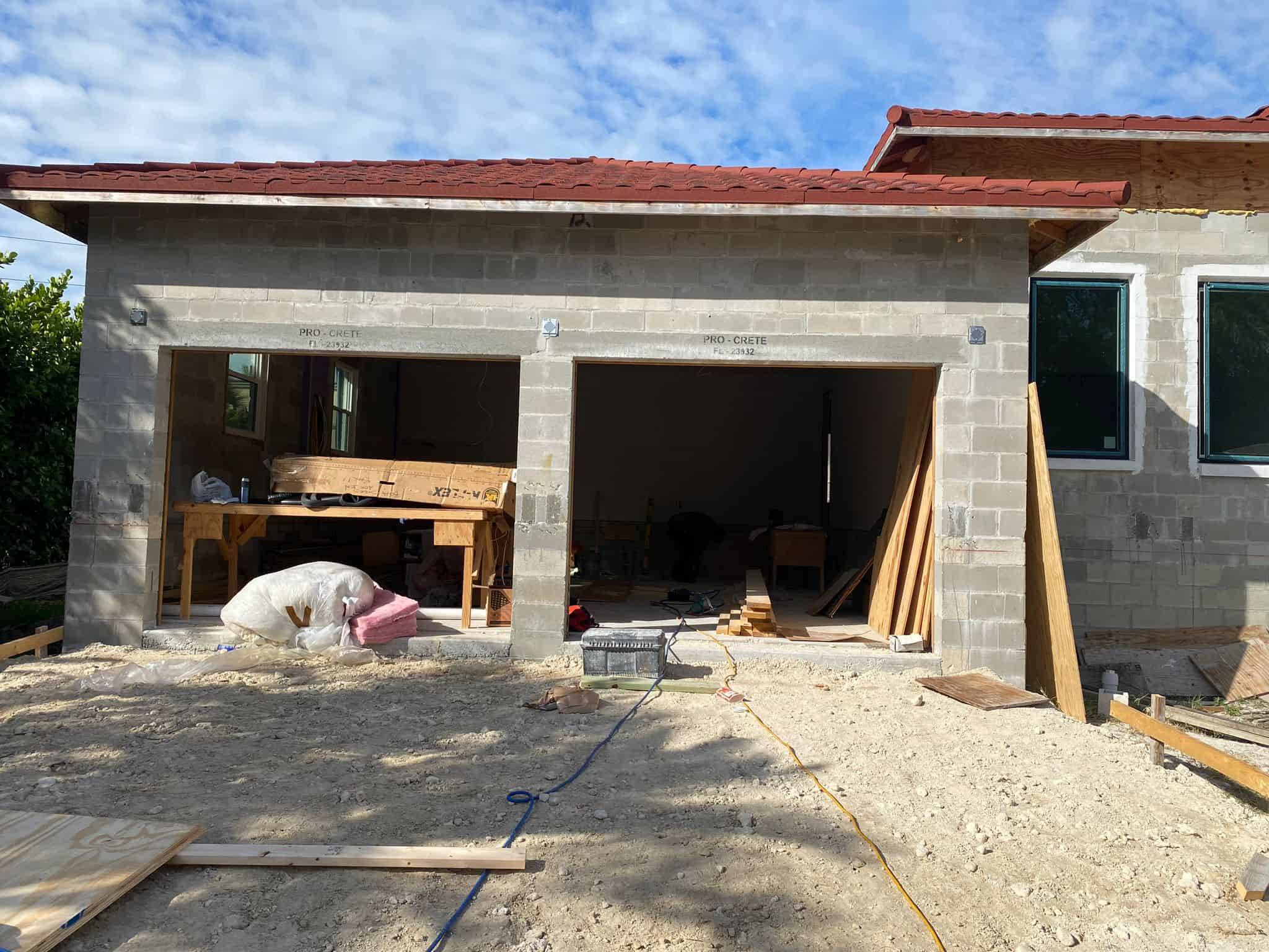 A partially constructed garage in Naples, Florida, with two large openings, building materials and tools scattered inside and outside, unfinished concrete block walls, and a red roof under a partly cloudy 34102 sky.