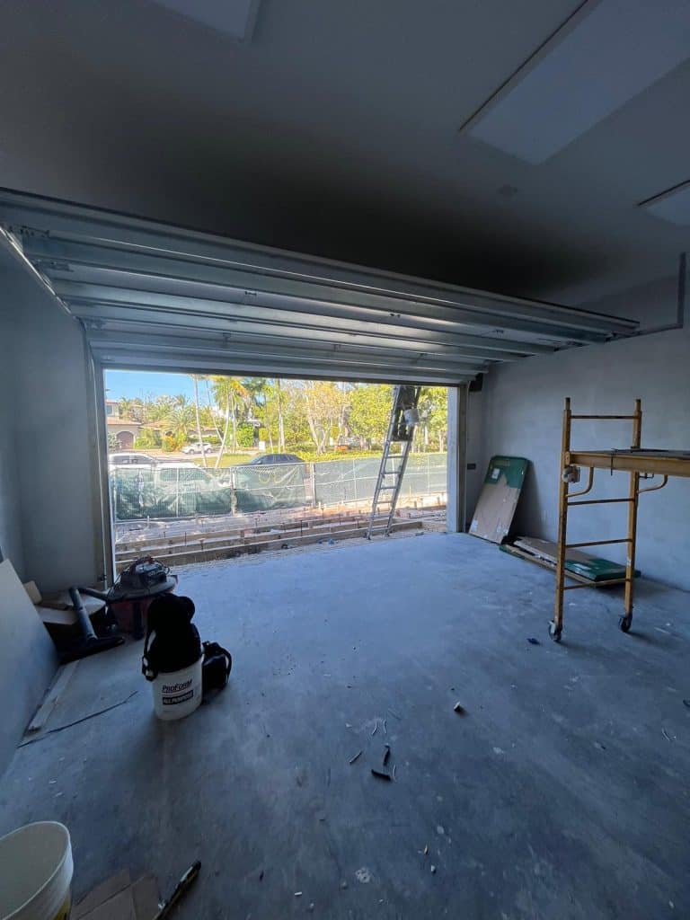 A garage under construction in Bay Harbor Islands, Florida, USA, with an open door, exposed ceiling tracks, a ladder outside, scaffolding inside, and construction tools scattered on the floor. Trees and a fence are visible in the background.