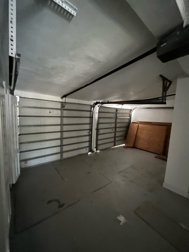 A dimly lit, empty garage in North Miami, Florida, with a closed sectional garage door, white walls, and a few boards leaning against the side wall. The floor has some stains and debris.