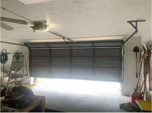 A partially open garage door lets in light. Tools and equipment are organized on shelves, hooks, and the floor along the walls. A ceiling fan hangs overhead, and garden tools are stored to the right in this Bonita Springs, Florida garage.
