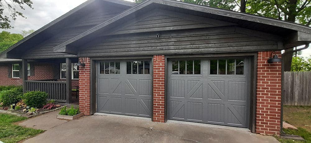 A brick house with a dark wooden roof and two gray residential garage doors. Window panels are at the top of each door. Plants and shrubs line the left side, with a wooden fence and trees in the background. Ideal for new garage door installation.