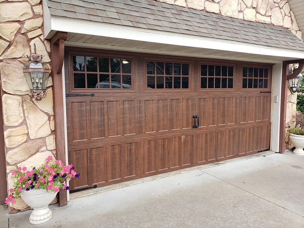 A brown wooden residential garage door with decorative black hardware and rectangular windows along the top, set in a stone exterior wall. Two wall lanterns and a white planter with pink flowers sit beside the driveway.