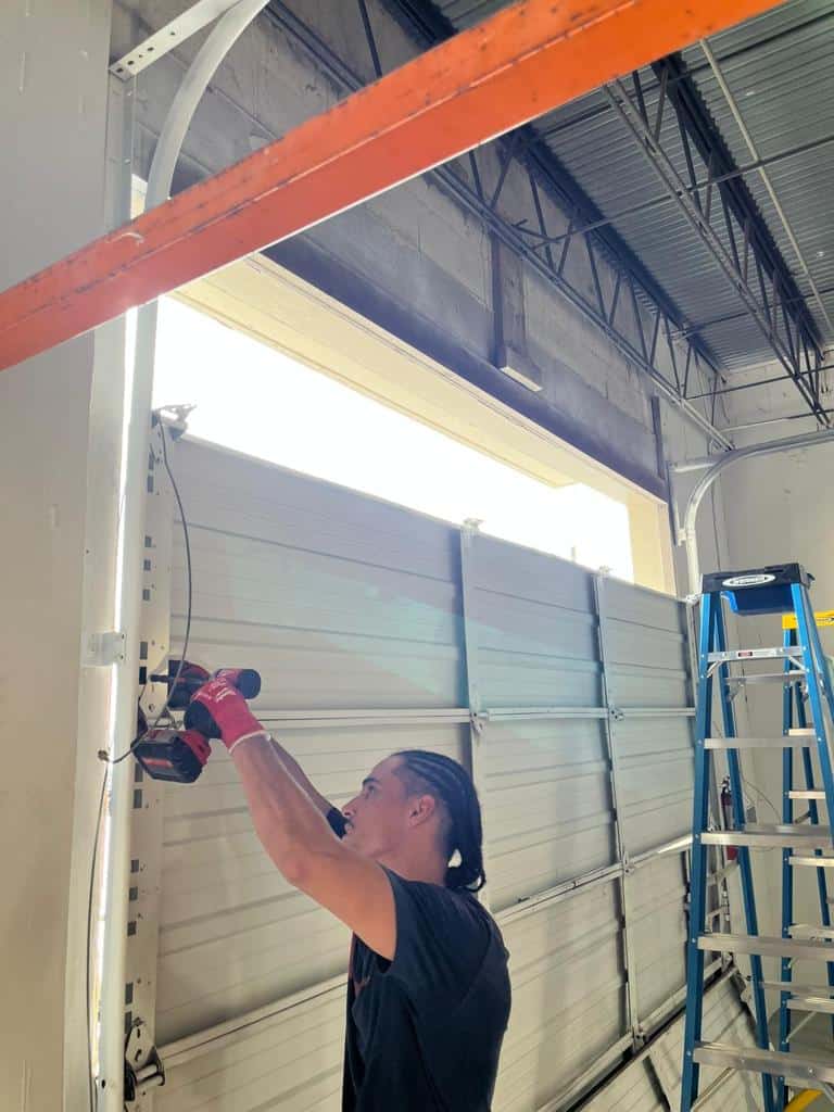 A person with braided hair uses a power drill to repair or install commercial garage doors in a workshop. A blue ladder is nearby, and sunlight streams through the upper window.