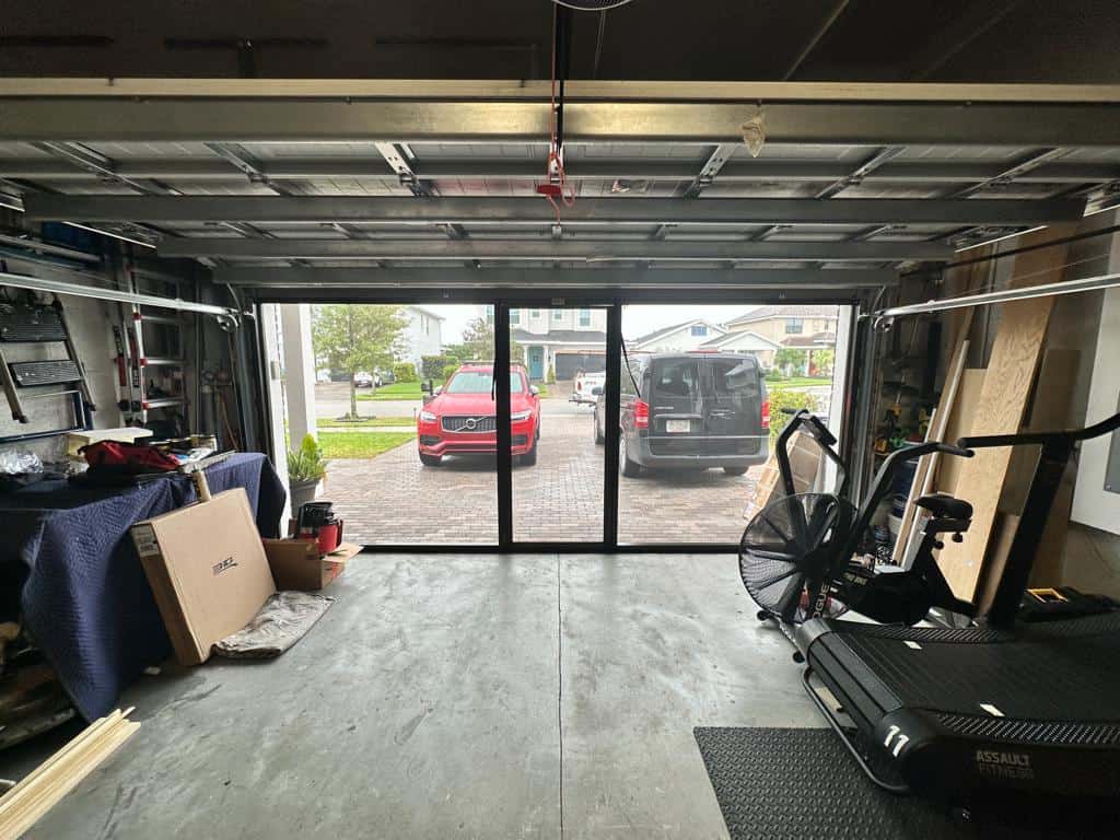 View from inside a garage with the door open, featuring Garage Door Screens, a red car and black van on the driveway. The garage holds exercise equipment, boxes, and various tools neatly arranged along the sides.