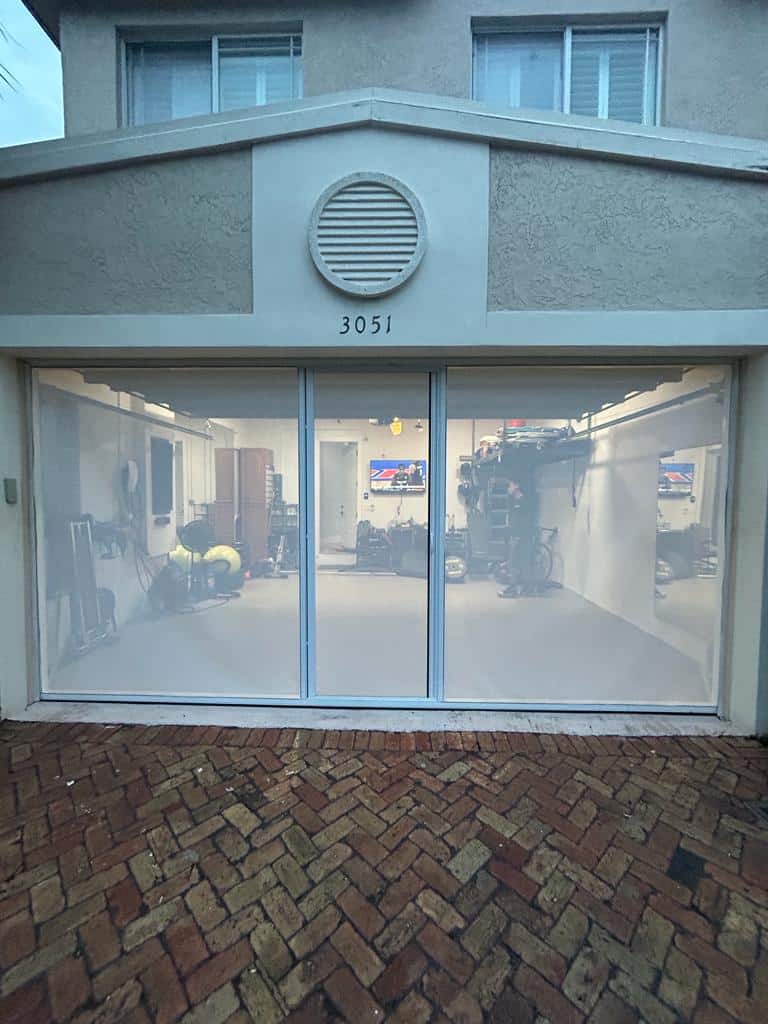 A garage with Garage Door Screens features a translucent screen door revealing exercise equipment and a TV inside. The address "3051" is above the entrance, and the wet brick driveway suggests recent rain.