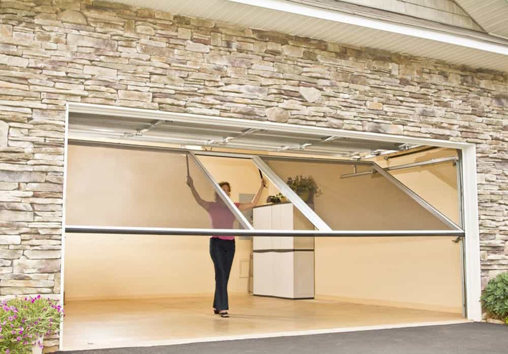 A woman opens a large garage door with a stone exterior. Inside, the space is empty except for a white storage cabinet in the back corner, accessorized with some plants nearby.