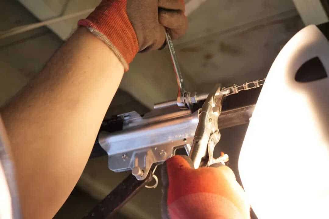In a dimly lit setting, a person wearing gloves skillfully uses a wrench and pliers to tighten a bolt on the metal mechanism during a garage door opener installation.