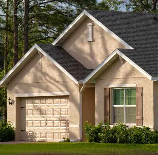 A suburban house with a beige exterior, dark roof, and a sleek garage door features eye-catching garage door accessories. It has a small front yard with green grass and shrubs, while trees create a charming backdrop.