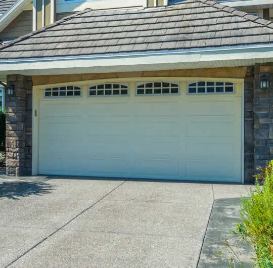 A two-car garage with a white sectional door and a garage door opener installation showcases three rectangular windows at the top. The house exterior combines stone and siding under a sloped roof, while the smooth concrete driveway leads up to this stylish feature.