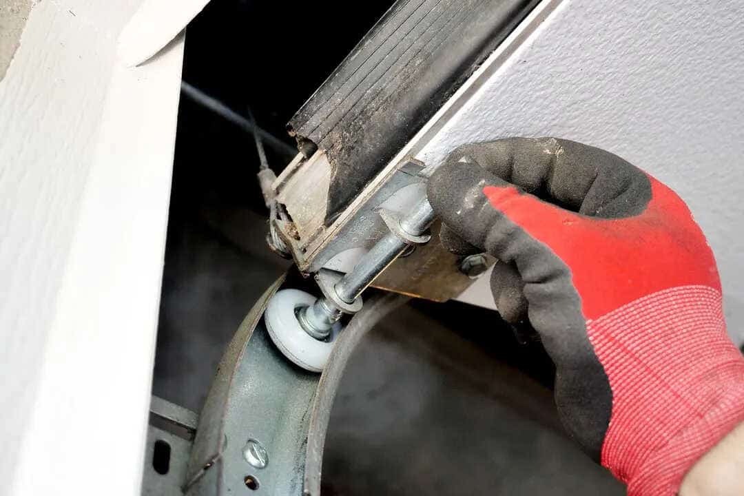 A person wearing a red and black glove is adjusting a metal bracket with a wheel on the garage door track, showcasing the intricacies of garage door installation. The focus is on the hand and components critical to the mechanism's smooth operation.