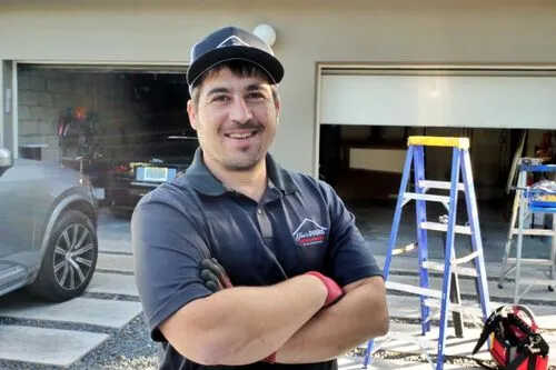 A smiling man in a navy blue polo shirt and cap stands with folded arms in front of a garage. Ladders and tools are visible behind him, suggesting he is working on a seal replacement or other home project.