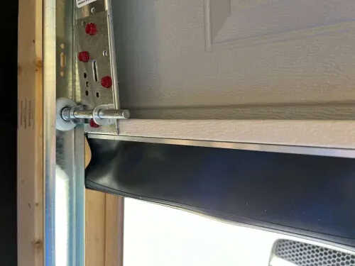 Close-up of a garage door's edge, focusing on the metal track, bolts, and black rubber weatherstripping seal during seal installation. Some red adjustment screws are visible on the metal bracket.