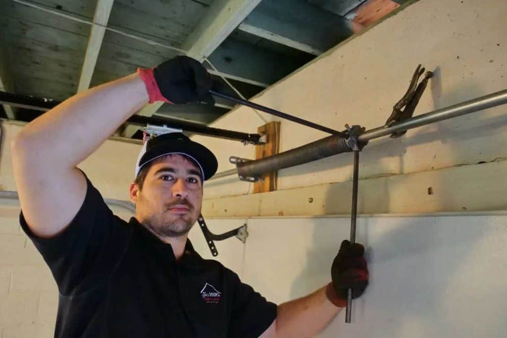 A person in a black shirt and hat is expertly working on a metal garage door mechanism using a wrench and a rod. Amidst exposed wooden planks, they focus intently under the beam, ensuring the garage door accessories are in perfect order for smooth operation.