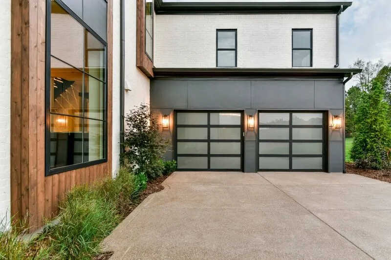 Modern house exterior with a two-car garage featuring frosted glass doors, showcasing recent garage door installation. Large windows are visible on the left, with wall-mounted lanterns flanking the garage. A paved driveway and neatly landscaped greenery complete the scene.
