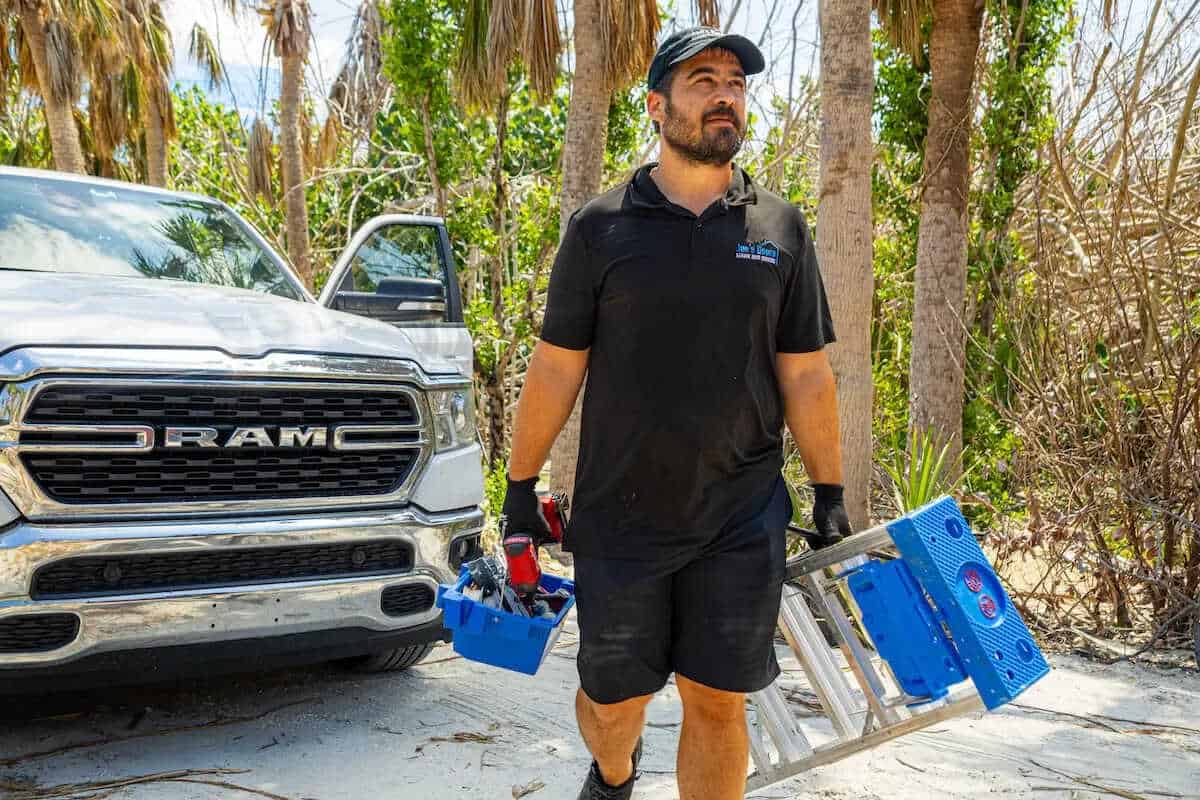 A man in a black polo and shorts carries a blue toolbox and a ladder, walking on a sandy surface beside an open RAM truck. Ready for action, he's likely en route to a garage door installation amid the backdrop of tropical plants and palm trees.