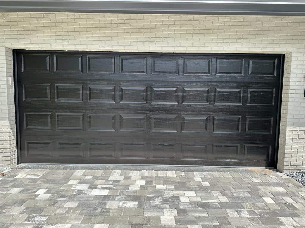 A black, paneled garage door set in a light beige brick wall, with a gray and white patterned stone driveway in front—showcasing the modern appeal and garage door benefits of style and security.