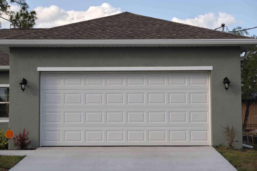A closed white garage door on a modern house with gray stucco exterior, two black outdoor wall lights on either side, and small shrubs along the concrete driveway showcase a property maintained to Better Business Bureau (BBB) standards.